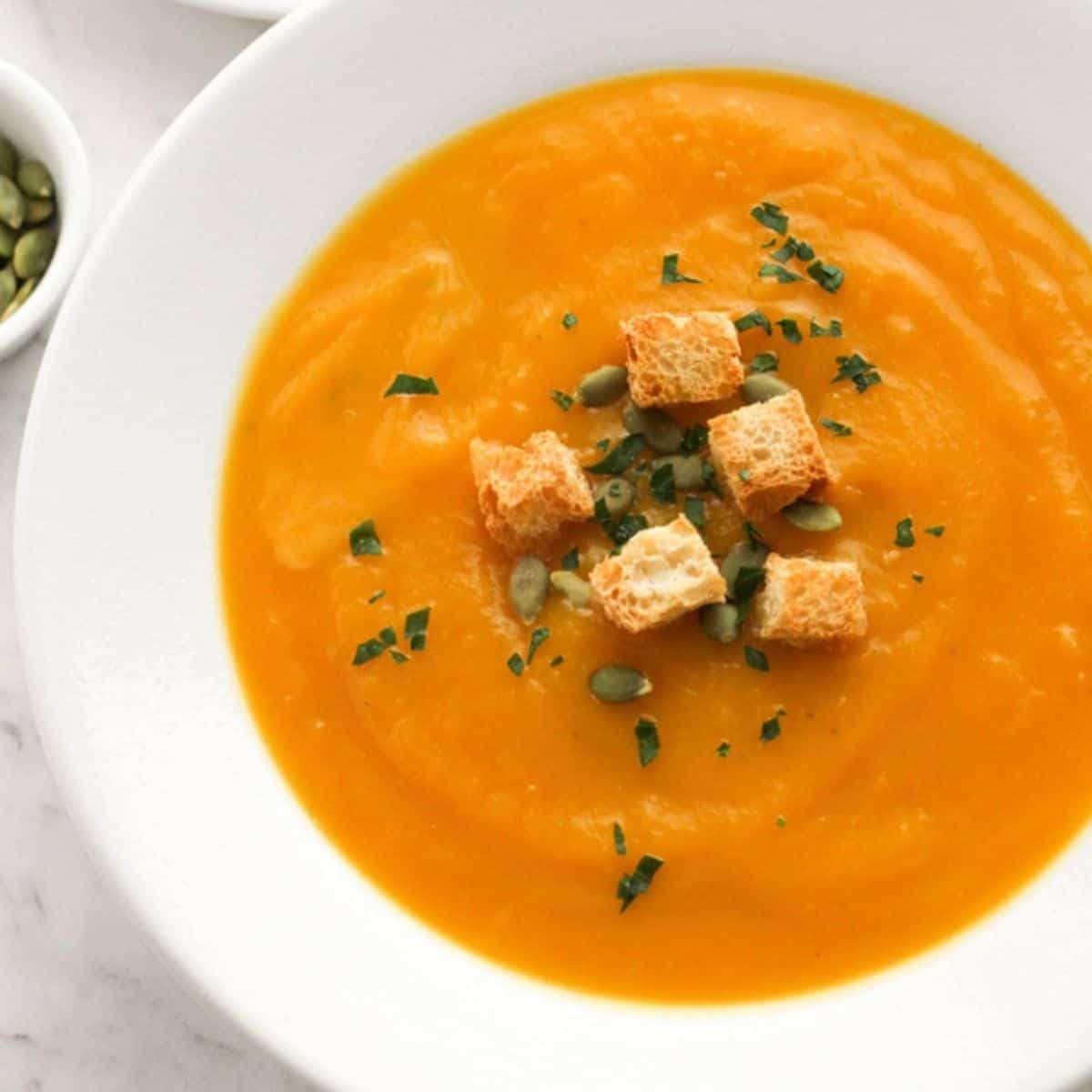 pumpkin soup in a white bowl topped with croutons and pumpkin seeds.
