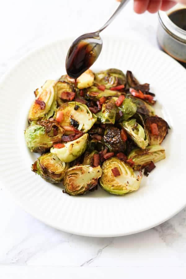 a spoon held above a plate of roasted brussels sprouts drizzling balsamic glaze over the vegetables