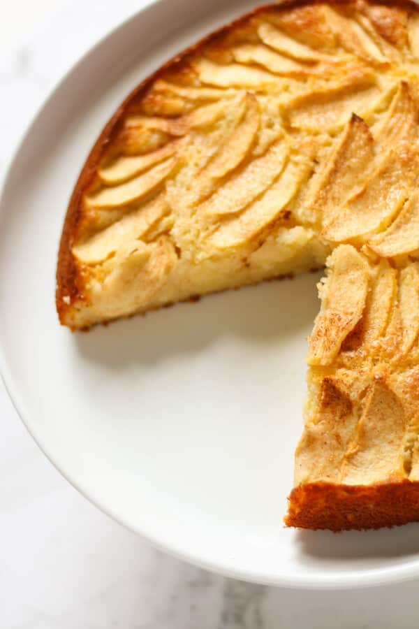 apple cake with a slice missing on a white cake stand.