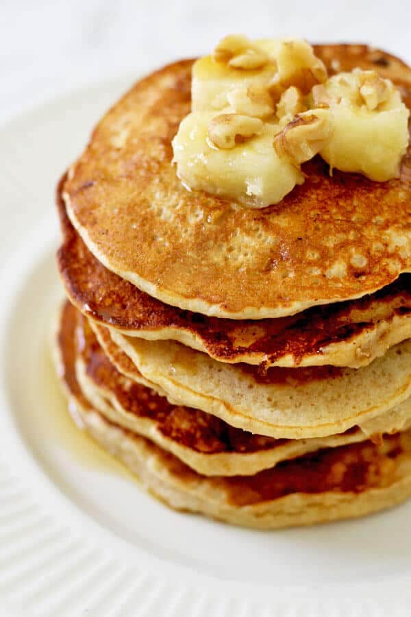 healthy banana pancakes stacked on a white plate with caramelised banana slices and pecans on top 