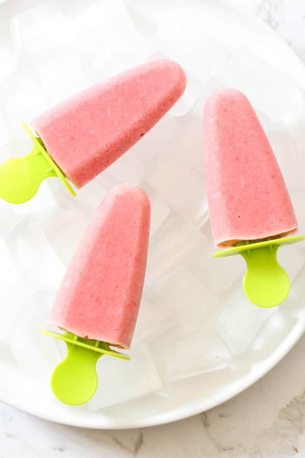3 strawberry pineapple popsicles on a plate filled with ice cubes.