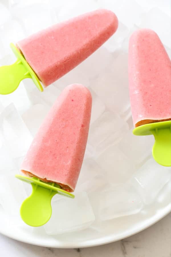3 strawberry pineapple popsicles on a plate filled with ice cubes.