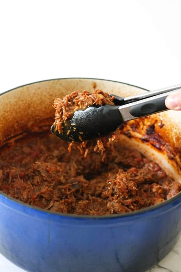 tongs pulling out shredded beef from blue dutch oven.