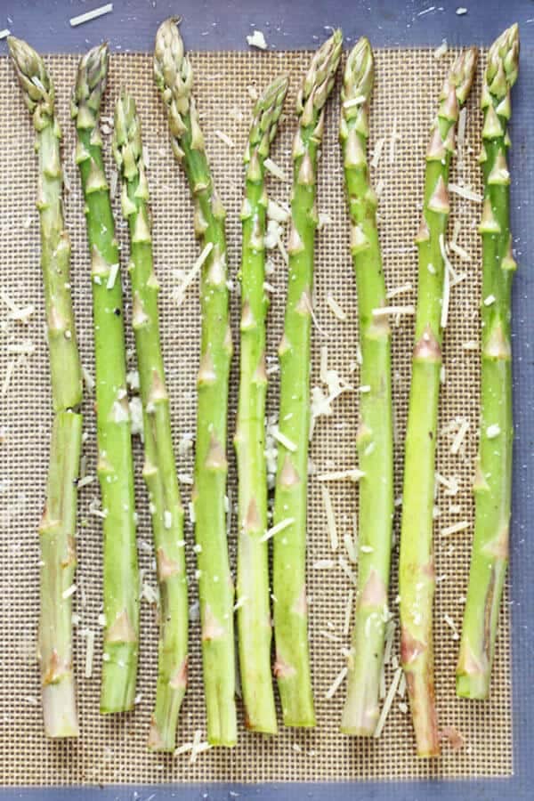 raw asparagus spears laid out in a single layer on a baking tray covered in parmesan cheese and minced garlic