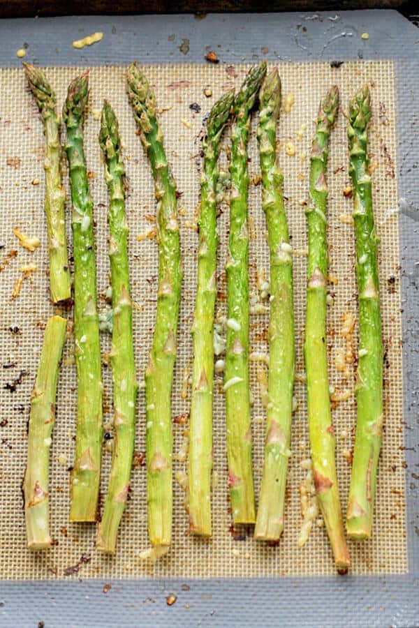cooked asparagus spears laid out in a single layer on a baking tray covered in parmesan cheese and minced garlic