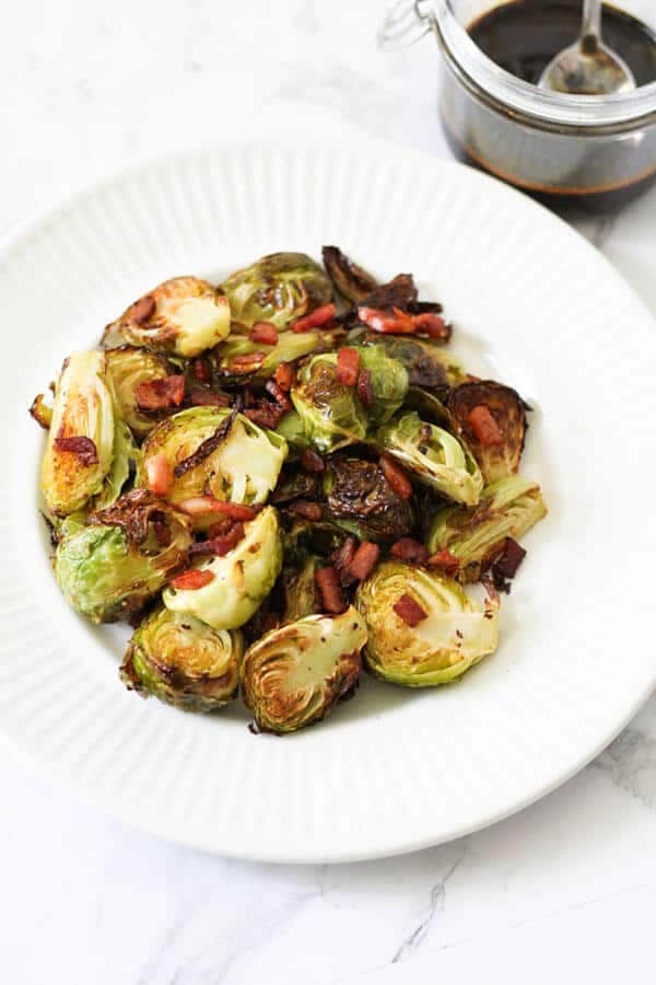 roasted brussels sprouts with bacon on a white plate with a jar of balsamic glaze in the background