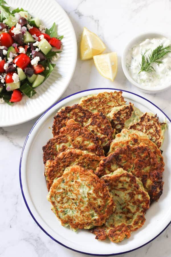 a plate of zucchini fritters flanked by a bowl of tzatziki and a plate of greek salad