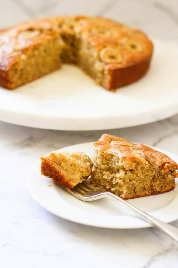 a slice of banana cake on a white plate with the rest of the cake on a wooden board behind.