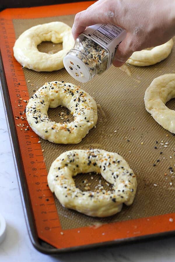 bagels on a baking tray being sprinkled with everything but the bagel seasoning. 