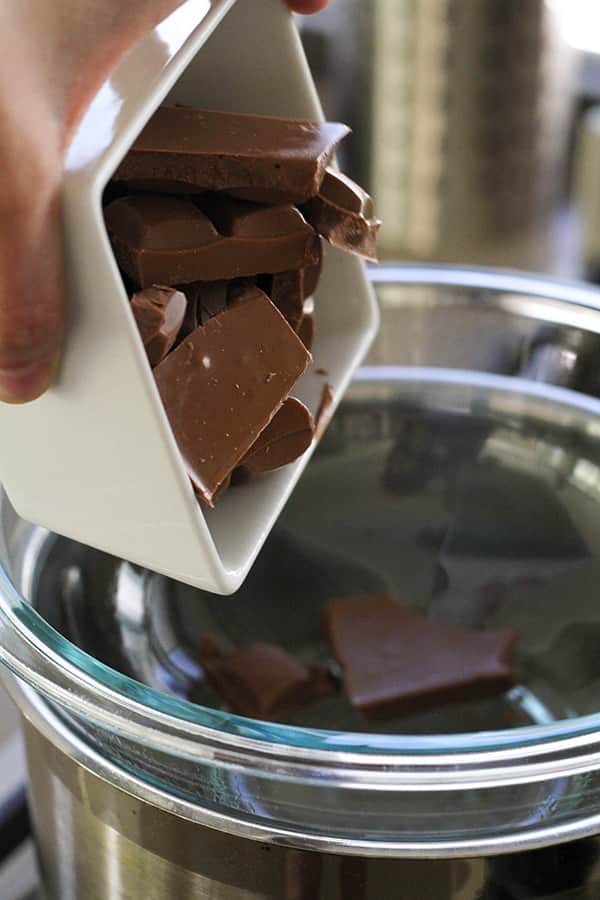 A hand pouring a bowl of chopped chocolate into a glass bowl over a saucepan.