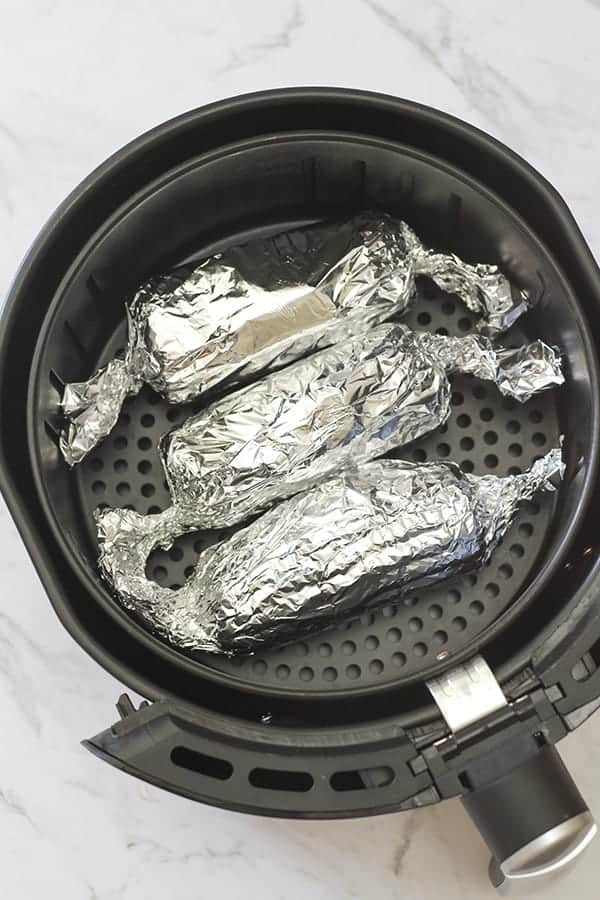 3 ears of corn wrapped in aluminium foil in an air fryer basket. 