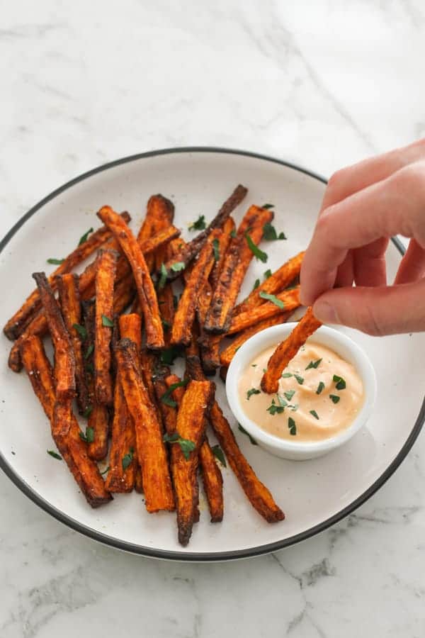 carrot fries on a white plate with a hand dipping a fry into sauce.