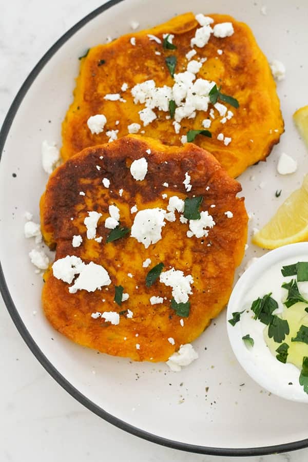 two pumpkin fritters on a white plate topped with feta and parsley.