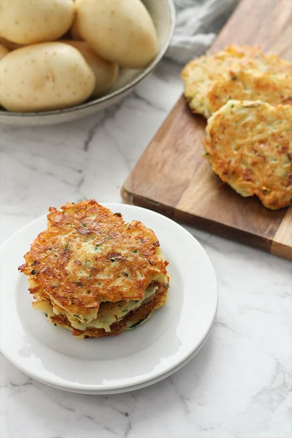 stack of potato fritters on a plate with a bowl of potatoes in the background.