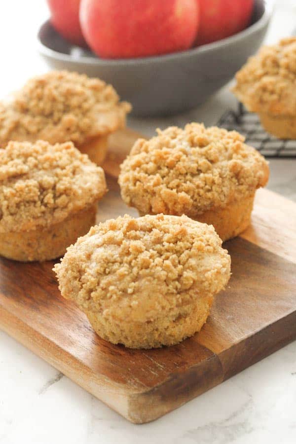 muffins on a wooden board with a bowl of apples in the background.
