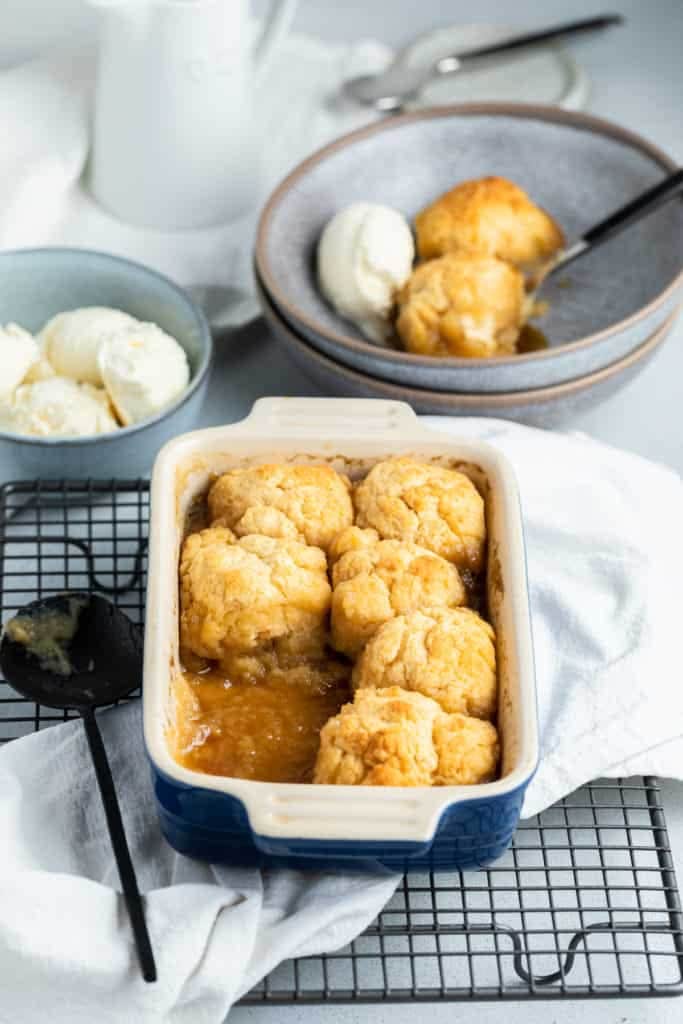 golden syrup dumplings in a baking dish on top of a wire tray with a bowl of ice cream in the background. 