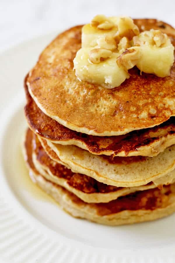 healthy banana pancakes stacked on a white plate with caramelised banana slices and pecans on top