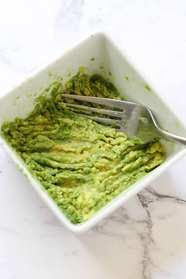 a fork pressing into mashed avocado in a white bowl