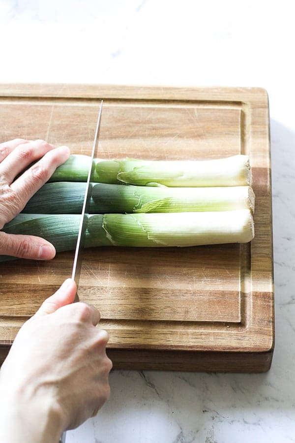 Leeks on a wooden chopping board with a hand cutting the dark ends off. 