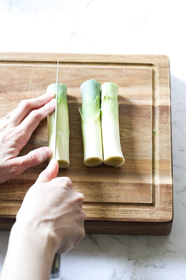 Leeks on a wooden chopping board with a hand slicing the leeks in half. 