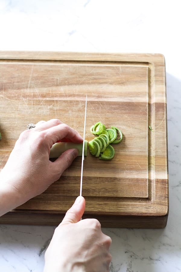 Leeks on a wooden chopping board with a hand slicing the leeks into half moons.