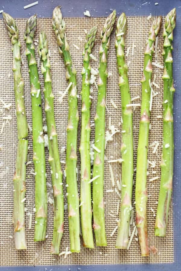 raw asparagus spears laid out in a single layer on a baking tray covered in parmesan cheese and minced garlic