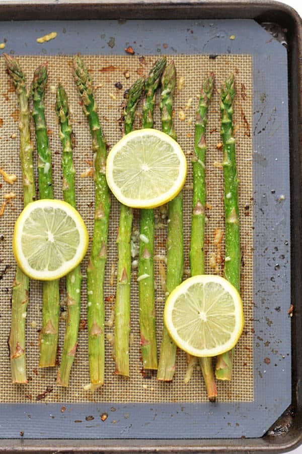 asparagus spears laid out in a single layer on a baking tray covered in parmesan cheese, minced garlic and lemon slices