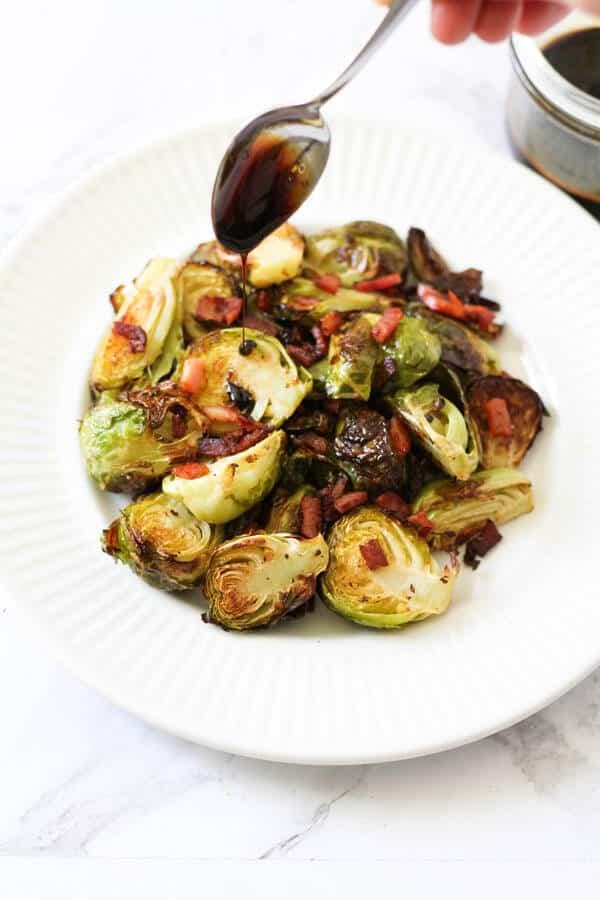 a spoon held above a plate of roasted brussels sprouts drizzling balsamic glaze over the vegetables