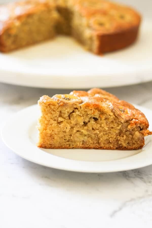 a slice of banana cake on a white plate with the rest of the cake on a wooden board behind.