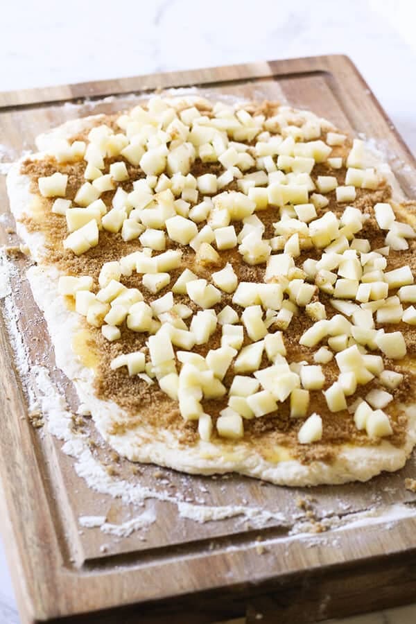 dough rolled out on a wooden board covered with sugar, cinnamon and apple slices. 