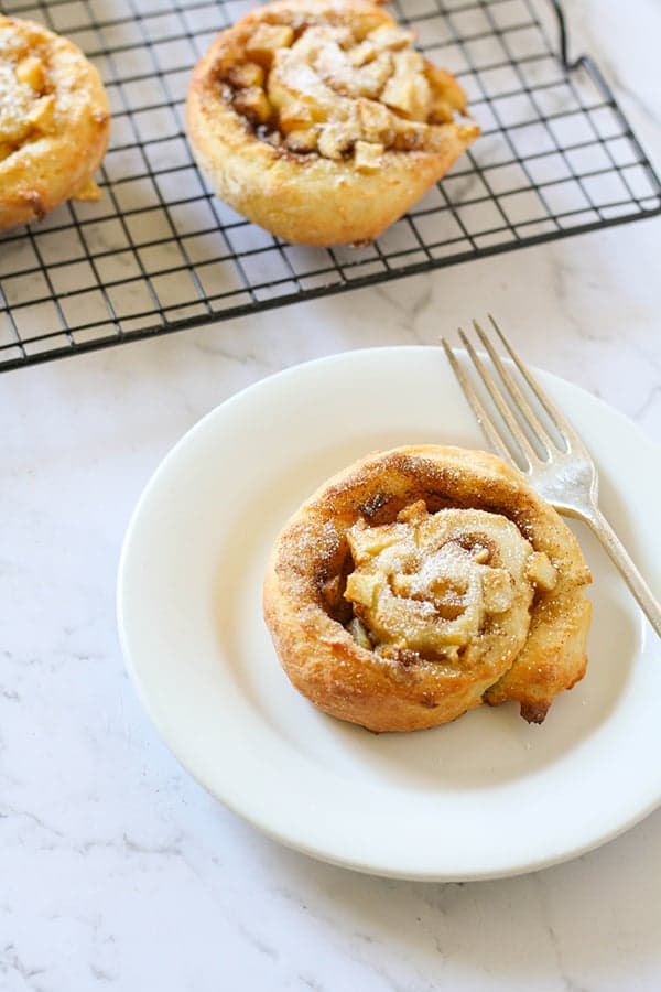 an apple cinnamon rolls on a white plate with a wire rack filled with rolls in the background.