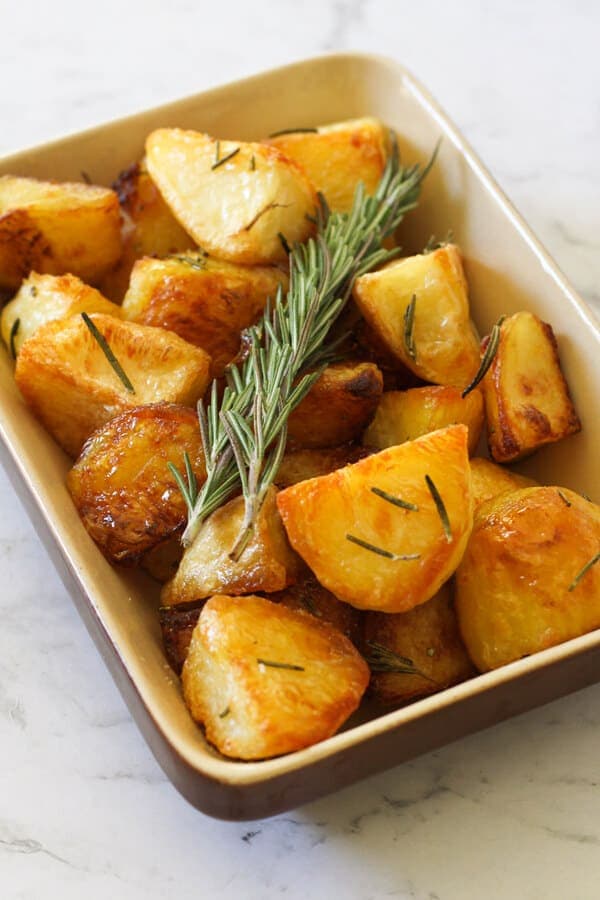 Crispy roast potatoes in a serving tray with rosemary sprigs on top.