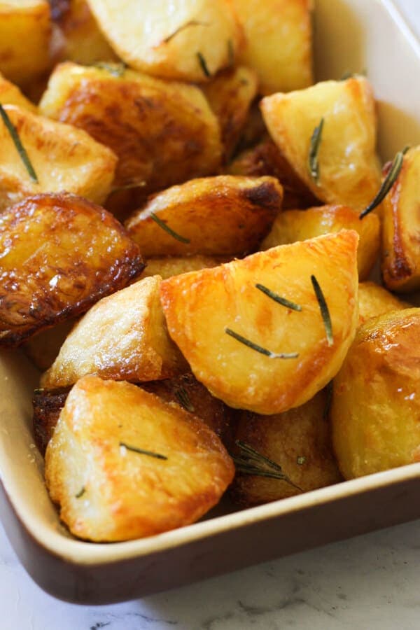 Crispy roast potatoes in a serving tray with rosemary sprigs on top.