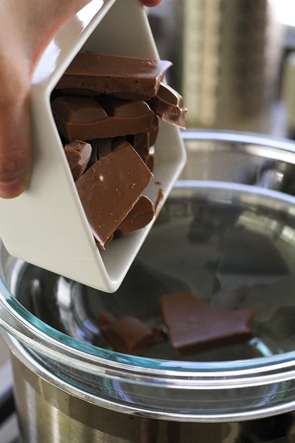 A hand pouring a bowl of chopped chocolate into a glass bowl over a saucepan. 