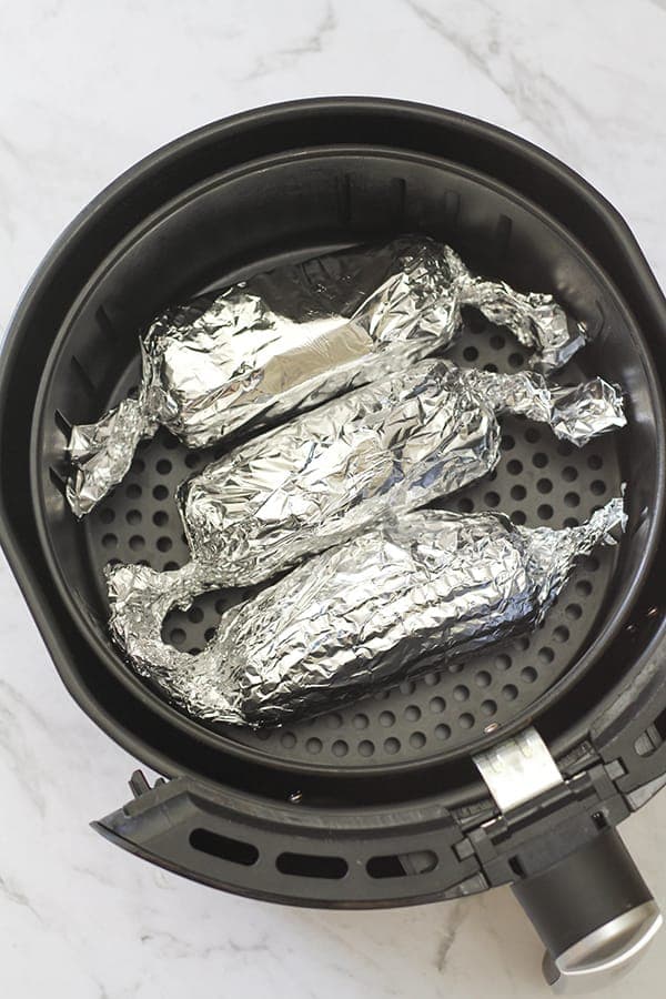3 ears of corn wrapped in aluminium foil in an air fryer basket. 