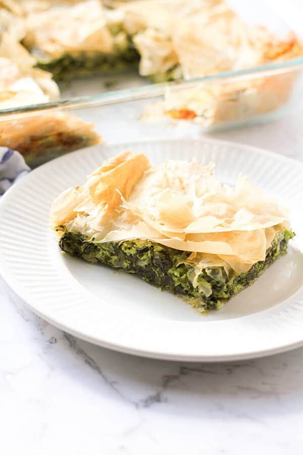 a slice of spanakopita on a white plate with a tray on spanakopita behind.