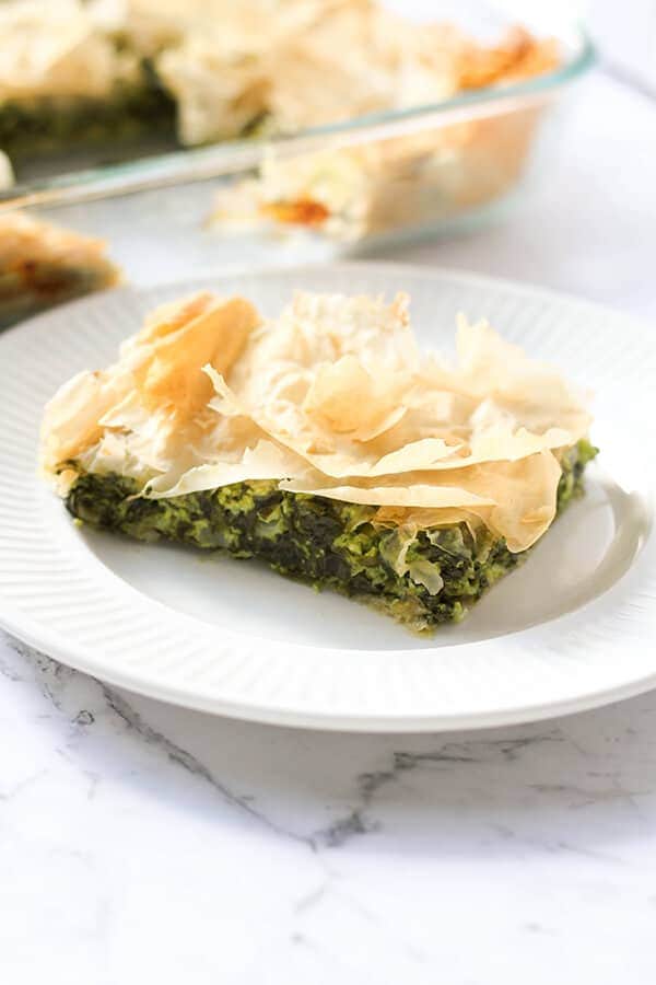 a slice of spanakopita on a white plate with a tray on spanakopita behind.