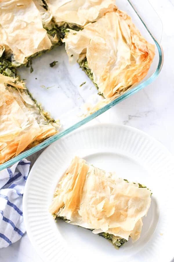 a slice of spanakopita on a white plate with a tray on spanakopita behind.