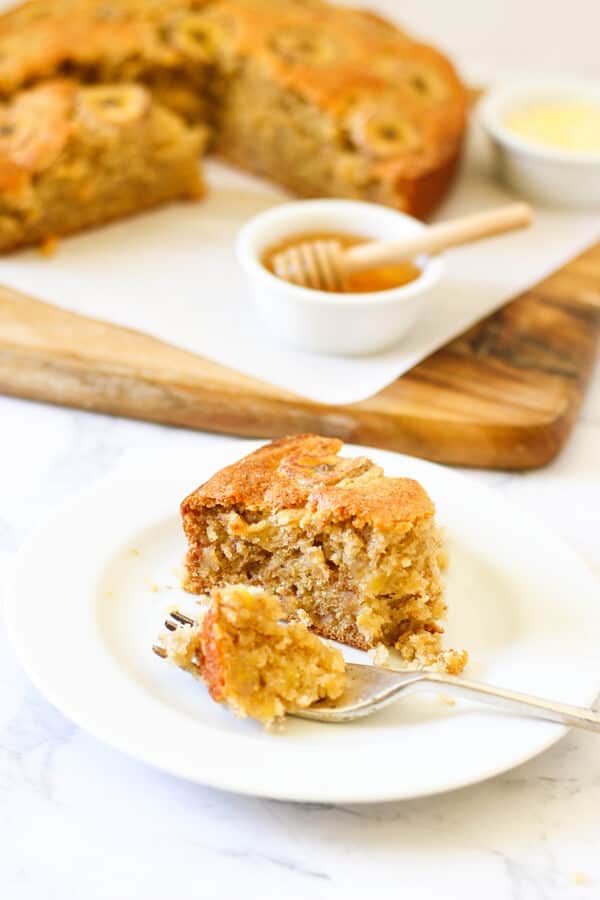a slice of banana cake on a white plate with the rest of the cake on a wooden board behind.